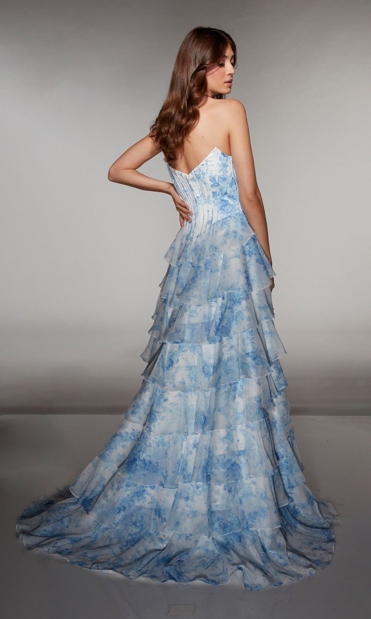 A woman poses with her back to the camera against a plain background, wearing the Alyce 62192 long prom dress with a strapless corset bodice and tiered blue and white patterned skirt.